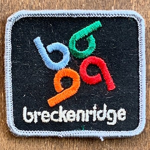 Op de afbeelding: Geborduurde patch met het woord "breckenridge" in het wit, onder kleurrijke, gestileerde cijfers. De cijfers zijn blauw, groen, rood en oranje, op een zwarte achtergrond met een grijze rand.