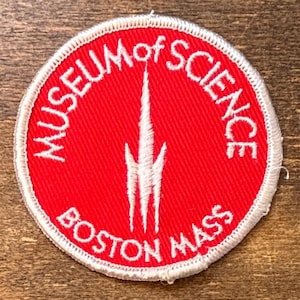 May include: A circular embroidered patch with a bright red background and a white border. The text "MUSEUM of SCIENCE" is arched across the top, and "BOSTON MASS" is arched across the bottom in white thread. A white stylized spire icon sits in the center.