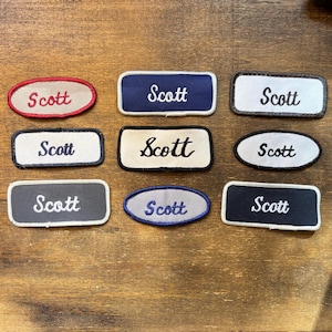 May include: A collection of nine embroidered patches, each featuring the name Scott in cursive script. The patches vary in shape, including rectangular and oval designs, and feature diverse color combinations such as red, blue, white, black, and grey.