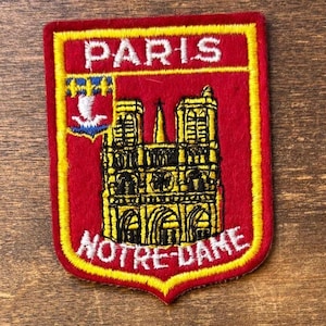 May include: A rectangular red embroidered patch with a yellow border featuring the white text PARIS at the top and NOTRE-DAME at the bottom. The center displays a yellow and black depiction of the Notre-Dame cathedral and a small blue shield.