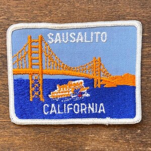 May include: Embroidered patch featuring the Golden Gate Bridge in Sausalito, California. The patch has a blue background with an orange bridge and a white border. The words "SAUSALITO" and "CALIFORNIA" are embroidered on the patch.