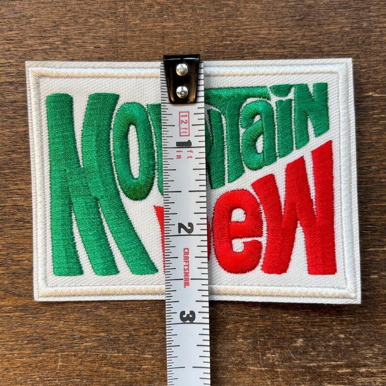 Op de afbeelding: Geborduurde patch met de woorden "Mountain Dew" in groen en rood. De patch is rechthoekig, ongeveer 6,35 cm hoog, met een witte rand en een cr&egrave;mekleurige achtergrond. De tekst is in een gestileerd lettertype.