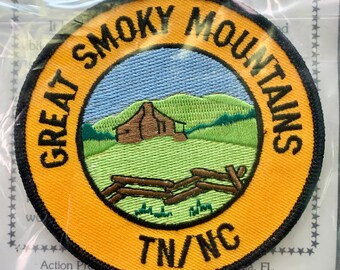 May include: A yellow and black embroidered patch with the text "Great Smoky Mountains TN/NC". The patch features a cabin in a green field with a blue sky and a wooden fence.
