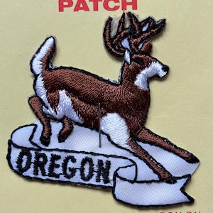 Puede incluir: Parche bordado marrón y blanco de un ciervo con cuernos de pie sobre una pancarta blanca con la palabra "OREGON" en negro.