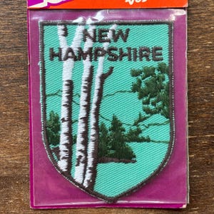 Puede incluir: Un parche bordado verde y marrón con el texto "NEW HAMPSHIRE" y un diseño de tres abedules frente a un bosque.
