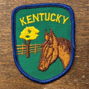 Puede incluir: Parche bordado con fondo verde y borde azul. El parche presenta la palabra "KENTUCKY" en amarillo, un árbol amarillo, una cabeza de caballo marrón y una valla marrón.