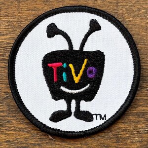 May include: A round, white patch with a black border featuring a black cartoon television character with antennae and legs. The word "TiVo" is written in colorful letters across the TV screen.