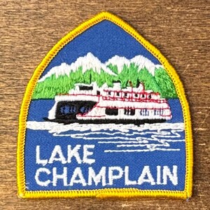 Op de afbeelding: Geborduurde patch met een veerboot op Lake Champlain, met bergen op de achtergrond. De patch is blauw met witte, rode, groene en gele accenten. De tekst op de patch luidt "LAKE CHAMPLAIN".