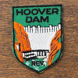 Può includere: Toppa ricamata raffigurante la diga di Hoover. La toppa ha uno sfondo verde con le parole "HOOVER DAM" in nero. La diga è raffigurata in bianco, con formazioni rocciose arancioni e marroni su entrambi i lati. La parola "NEV" è in basso.