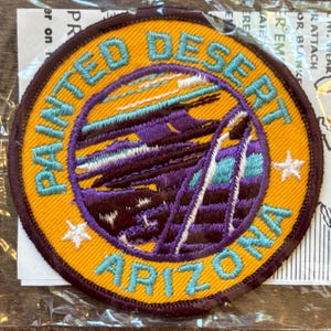May include: A round embroidered patch with the words "Painted Desert Arizona" in turquoise lettering. The patch features a desert landscape in purple, teal, and brown, with two white stars and a yellow border.