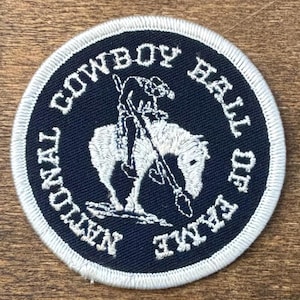May include: Round patch with a navy blue background and white embroidered text and design. The text reads "NATIONAL COWBOY HALL OF FAME". The design features a cowboy riding a horse. The patch has a white border.