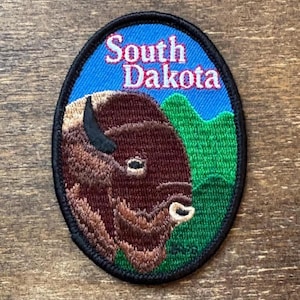 Könnte beinhalten: Ovaler Aufnäher mit einem Bison und den Worten "South Dakota" in weißer und roter Schrift auf blauem Hintergrund. Der Bison ist braun mit einem schwarzen Horn, und grüne Hügel sind rechts.
