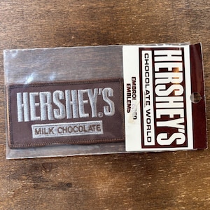 May include: A brown fabric patch with white embroidered text that reads "HERSHEY'S MILK CHOCOLATE". The patch is packaged in a clear plastic bag with a brown paper backing that reads "HERSHEY'S CHOCOLATE WORLD EMBROID. EMBLEMS".