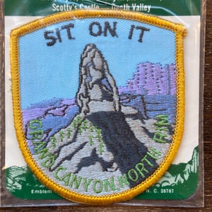 Op de afbeelding: Een geborduurde patch in geel en groen met de tekst "Sit on it" en een afbeelding van een rotsformatie. De tekst "Grand Canyon North Rim" is ook op de patch geborduurd.