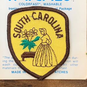 Op de afbeelding: Een gele viltpatch met een bruine rand in de vorm van een schild. De patch heeft de tekst "SOUTH CAROLINA" in een bruin cursief schrift rond een tekening van een vrouw in een bruine jurk die naast een groene plant met bruine bloemen staat.
