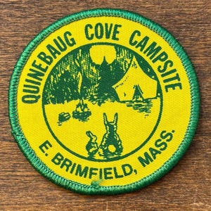 Könnte beinhalten: Ein runder, gelb-grüner Aufnäher mit dem Text "QUINEBAUG COVE CAMPSITE E. BRIMFIELD, MASS.". Der Aufnäher zeigt eine grüne Illustration eines Zeltes, Bäume und zwei Kaninchen.