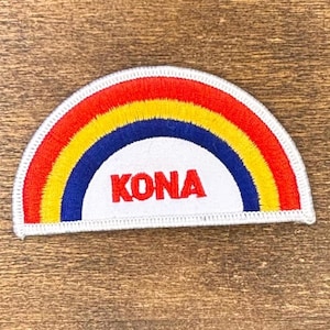 May include: Embroidered patch in a rainbow design with red, yellow, and blue stripes. The word "KONA" is embroidered in red on a white background. The patch is bordered with white stitching.