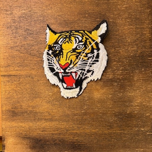 May include: An embroidered patch featuring the head of a tiger with yellow, black, and white fur. The tiger has an open mouth showing white teeth, a bright red tongue, and detailed black stripes across its face, ears, and whiskers.