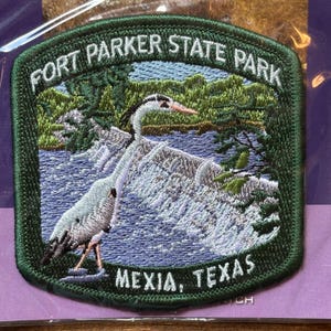 May include: Embroidered patch featuring a heron in a natural setting with water and trees. The patch is green with the text "FORT PARKER STATE PARK" and "MEXIA, TEXAS". The heron is gray and white with a yellow beak.