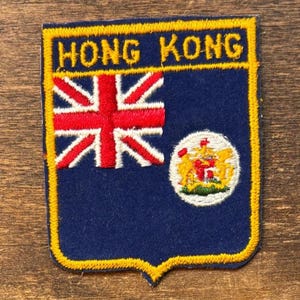 May include: A blue, shield-shaped patch with the words "HONG KONG" in yellow. The patch features the Union Jack flag and a circular emblem with a coat of arms. The patch is outlined in yellow.