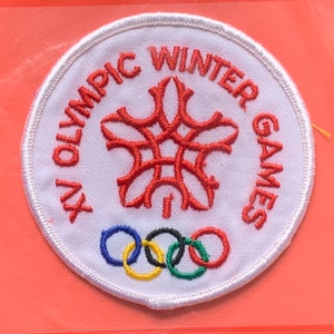 Könnte beinhalten: Ein weißer runder Patch mit rotem gesticktem Text, der "XV Olympic Winter Games" lautet. In der Mitte des Patches befindet sich eine rote stilisierte Schneeflocke. Fünf ineinandergreifende Ringe in Blau, Gelb, Schwarz, Grün und Rot befinden sich am unteren Rand des Patches.