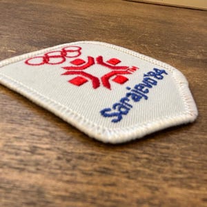 Puede incluir: Parche blanco bordado con detalles rojos y azules. El dise&ntilde;o incluye los anillos ol&iacute;mpicos, un emblema estilizado y el texto "Sarajevo 84" en azul. El parche tiene un borde blanco.