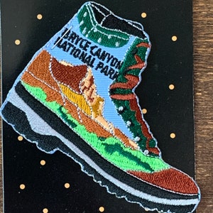 Puede incluir: Parche bordado con un diseño de bota de senderismo con el texto "Bryce Canyon National Park". La bota tiene una combinación de colores azul, marrón, verde y negro, con un borde blanco. El parche está sobre un fondo negro con puntos dorados.
