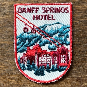 May include: Embroidered patch featuring the Banff Springs Hotel. The patch has a white background with red and teal accents. The text "BANFF SPRINGS HOTEL" is at the top. The design includes a hotel, trees, and a ski lift.