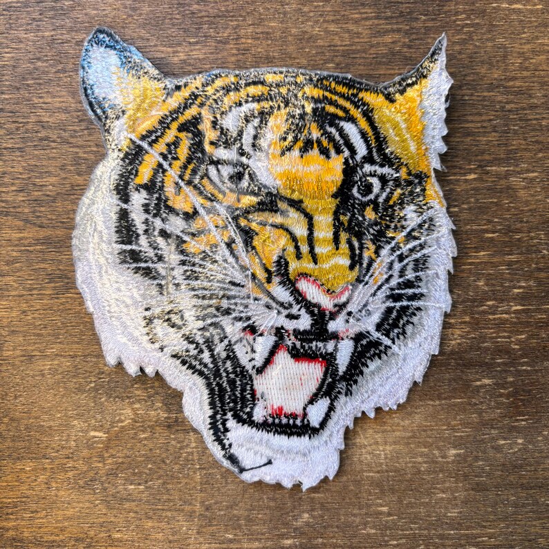 May include: An embroidered patch featuring the head of a tiger with yellow, black, and white thread. The tiger is depicted with an open mouth showing white teeth, a pink tongue, and detailed black stripes across its face and ears.