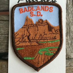 May include: Brown embroidered patch with a blue background. The patch features a landscape of brown mountains with green vegetation and the text "BADLANDS S.D."