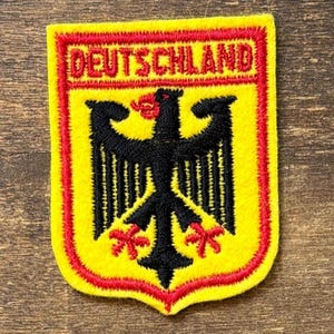 May include: A shield-shaped embroidered patch featuring a yellow background with a red border. The word DEUTSCHLAND is stitched in red at the top. A black eagle with red claws and a red beak is centered on the patch, mimicking the German coat of arms.