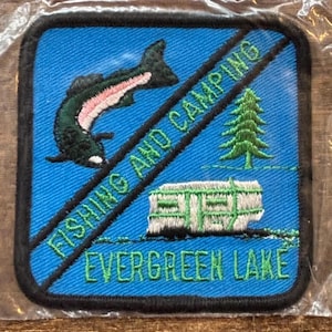 Op de afbeelding: Vierkante patch met een blauwe achtergrond, met een groene en roze vis, een groene dennenboom en een witte en groene camper. De patch leest "FISHING AND CAMPING" en "EVERGREEN LAKE" in groene letters.