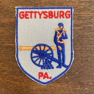 Könnte beinhalten: Ein weißer gestickter Aufnäher mit blauen und orangenen Details. Der Aufnäher zeigt eine Kanone und einen Soldaten mit dem Text "GETTYSBURG PA."