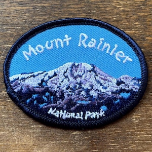 Puede incluir: Parche ovalado con un paisaje de montaña y el texto "Mount Rainier National Park". El parche tiene un fondo azul con una cordillera en tonos morados y blancos. El texto es blanco.