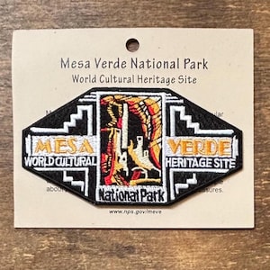 May include: A black and white patch with the text "Mesa Verde National Park World Cultural Heritage Site." The patch has a diamond shape with a colorful illustration of a building and the words "Mesa Verde" and "National Park."