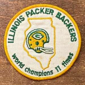 May include: A circular embroidered patch with a white background and a yellow border. It features a green outline of the state of Illinois containing a yellow and green football helmet. The text reads ILLINOIS PACKER BACKERS and WORLD CHAMPIONS 11 TIMES.