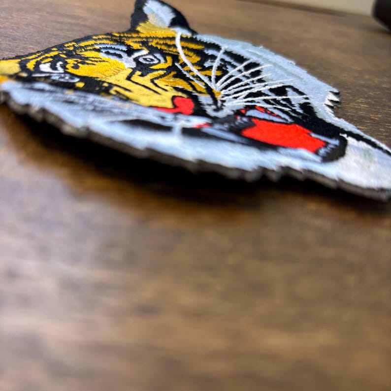 May include: An embroidered patch featuring the head of a tiger in profile. The design includes yellow, black, and white thread with a prominent red mouth and white whiskers. The patch has a light blue border and is shown from an angled perspective.