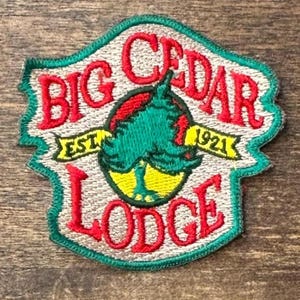 May include: An embroidered patch with a beige background and a green border. The text reads BIG CEDAR LODGE in red, with EST. 1921 in black on a yellow banner. A central circular graphic features a green tree silhouette against a red and yellow sun.