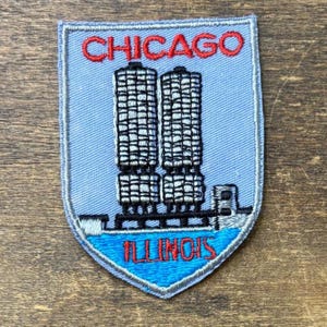 Puede incluir: Un parche bordado en forma de escudo con fondo azul claro. La palabra CHICAGO está bordada en rojo en la parte superior. Debajo, dos edificios cilíndricos grises se muestran sobre una sección de agua azul con la palabra ILLINOIS en rojo.