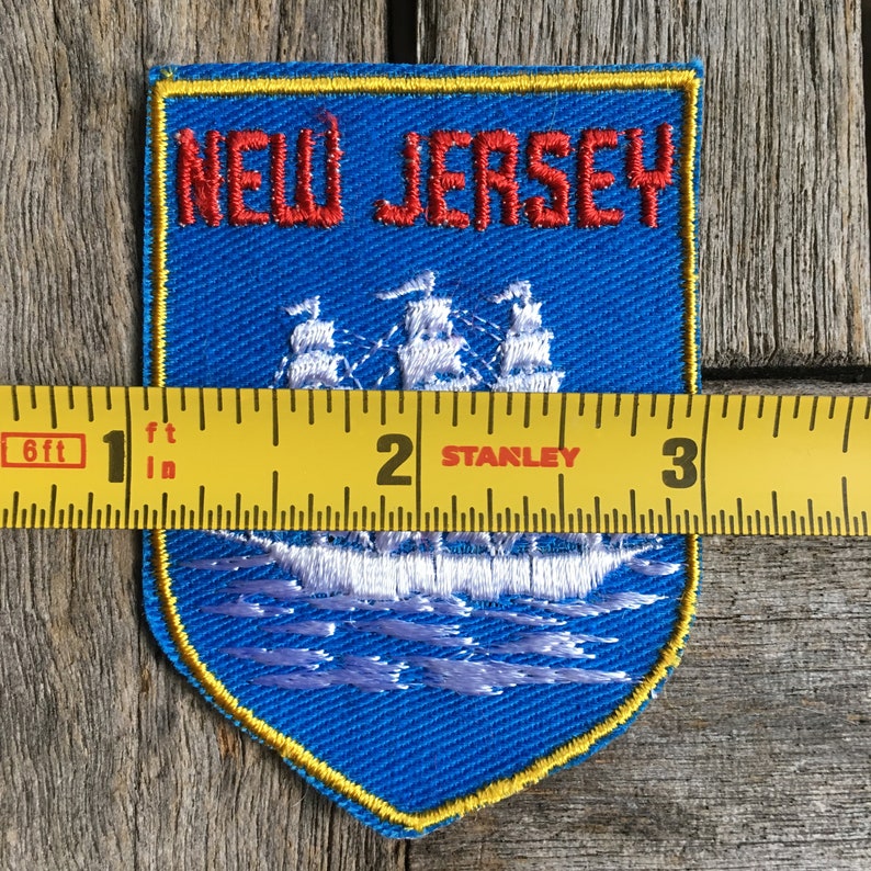 May include: A blue shield-shaped embroidered patch featuring the red text NEW JERSEY above a white sailing ship on water. A yellow Stanley measuring tape is placed across the patch, showing a width of approximately 8.25 cm.