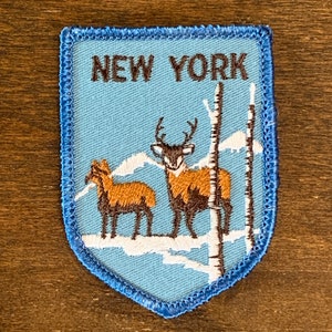 Könnte beinhalten: Gestickter Aufnäher mit dem Text "NEW YORK" über einer Szene mit zwei Hirschen, Schnee und Bäumen. Der Aufnäher hat einen hellblauen Hintergrund und einen blau genähten Rand.