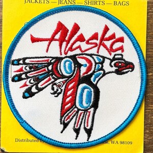 May include: Embroidered patch featuring the word "Alaska" in red script above a stylized bear design. The bear is depicted in black, red, and blue, with a white background and a blue border. The patch is on a yellow background.