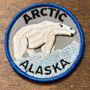 May include: Embroidered patch featuring a polar bear standing on ice. The patch has a gray background with the words "ARCTIC" and "ALASKA" in black lettering. A blue border surrounds the circular design.