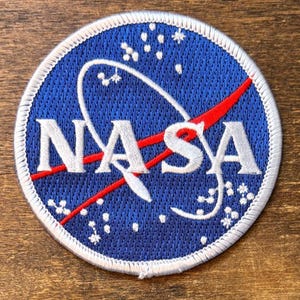 Könnte beinhalten: Gestickter NASA-Aufnäher mit dem Logo der Agentur. Der Aufnäher hat einen blauen Hintergrund mit weißem Text und einem roten Schwung. Das NASA-Logo ist von einem weißen Rand und kleinen weißen Sternen umgeben.