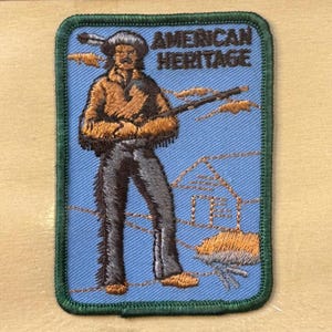 Könnte beinhalten: Gestickter Aufnäher mit blauem Hintergrund und grünem Rand. Der Aufnäher zeigt eine Figur in brauner und schwarzer Kleidung mit dem Text "AMERICAN HERITAGE" oben. Ein Haus und ein Baum sind ebenfalls abgebildet.