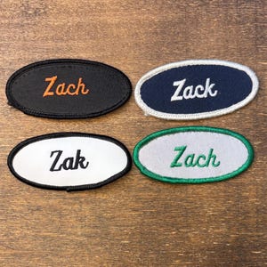 Puede incluir: Cuatro parches bordados de forma ovalada con bordes cosidos. Un parche negro con texto naranja que dice Zach, uno azul marino con texto blanco que dice Zack, uno blanco con texto negro que dice Zak y uno gris con texto verde que dice Zach.