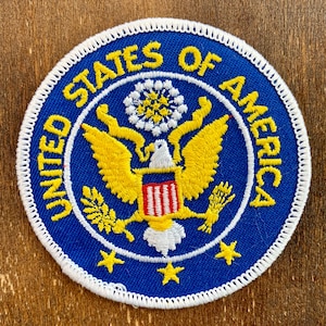 May include: A blue and white embroidered patch with the words "United States of America" surrounding the Great Seal of the United States. The seal features a bald eagle with outstretched wings, holding a bundle of arrows in one talon and an olive branch in the other. The eagle is perched on a shield with red and white stripes.