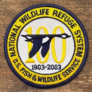 Puede incluir: Parche bordado circular con borde azul marino y texto amarillo. El parche dice "NATIONAL WILDLIFE REFUGE SYSTEM" y "U.S. FISH & WILDLIFE SERVICE". Un pájaro vuela sobre el número "100" y los años "1903-2003". El diseño tiene un fondo crema.