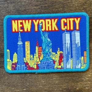 May include: A rectangular patch with a blue background and a turquoise border. The patch features the words "NEW YORK CITY" in yellow block letters. The image depicts the Statue of Liberty, the Twin Towers, and other New York City buildings.