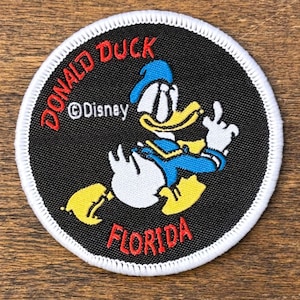 May include: Circular patch with a black background and white stitched edge, featuring Donald Duck. The text "DONALD DUCK FLORIDA" is in red, with "©Disney" in white. Donald Duck is wearing his blue sailor suit and a blue hat.